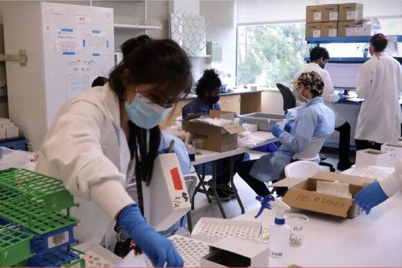scientist in gloves and mask working at a lab table within a larger lab